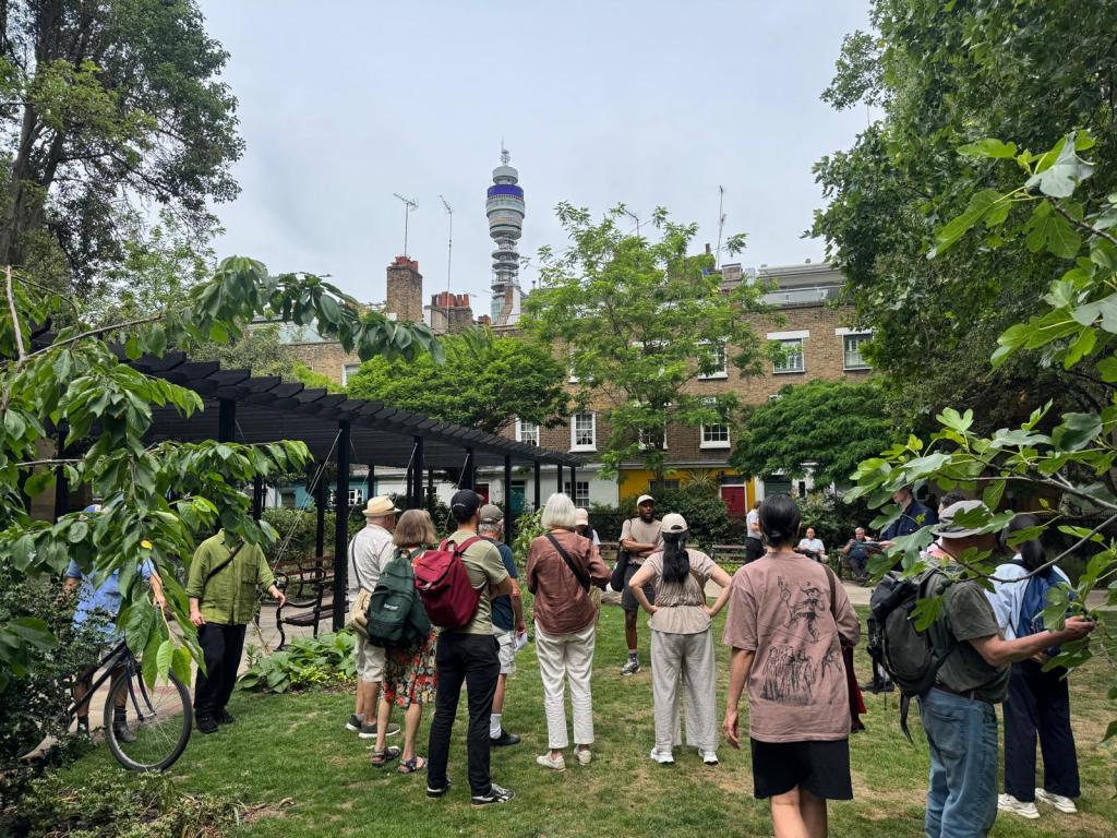 Group of people standing in a park with tree cover. The BT Tower is visible in the background above other buildings.